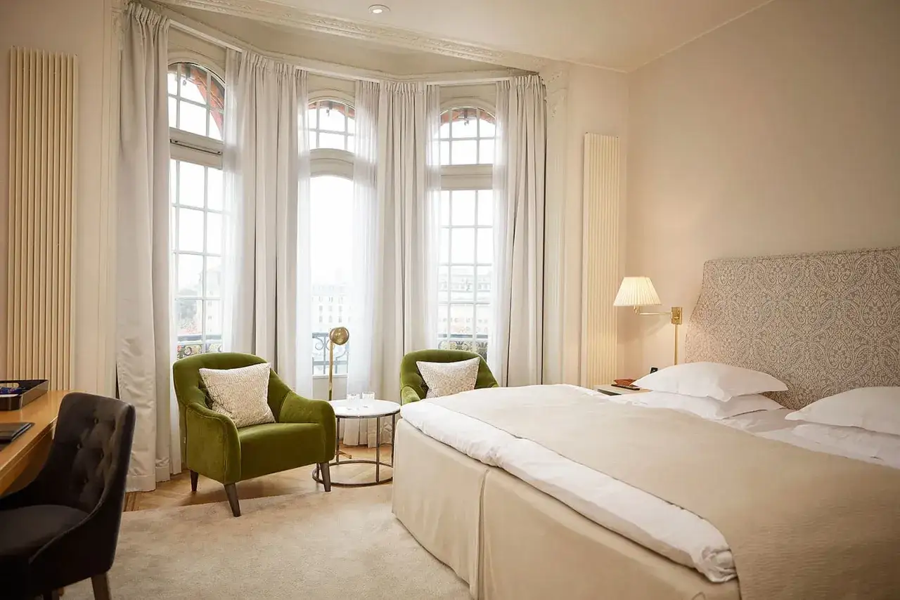 Ett elegant rum på Clarion Hotel Östermalm med en stor säng, gröna fåtöljer och vackra fönster med utsikt.