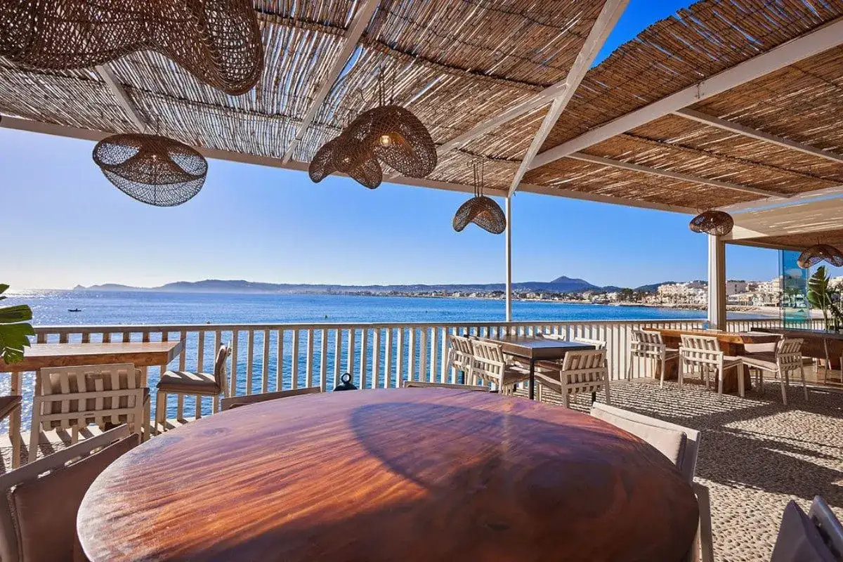 Terraza de restaurante con vistas al mar, mesas de madera y sillas, bajo un techo de paja. Un lugar perfecto para disfrutar en la colonia de carmela.