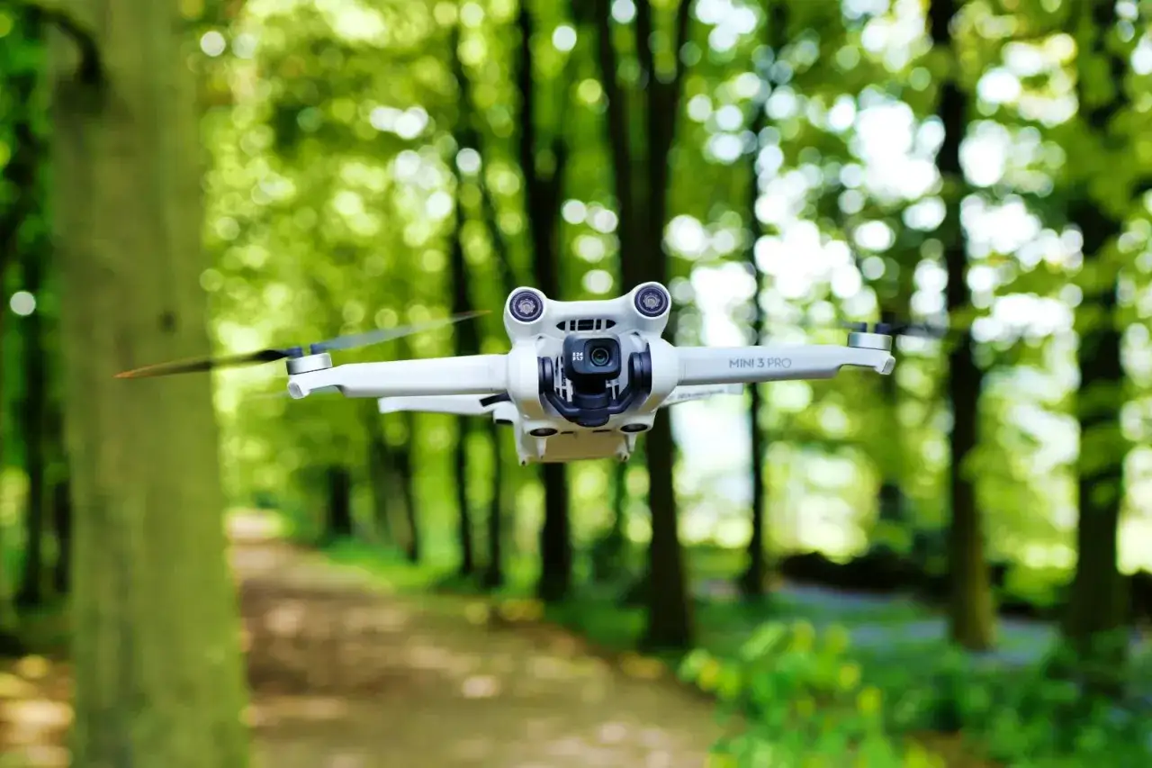 Biały dron DJI Mini 3 Pro w locie, na tle zielonego lasu. Jego zasięg pozwala na eksplorację natury.