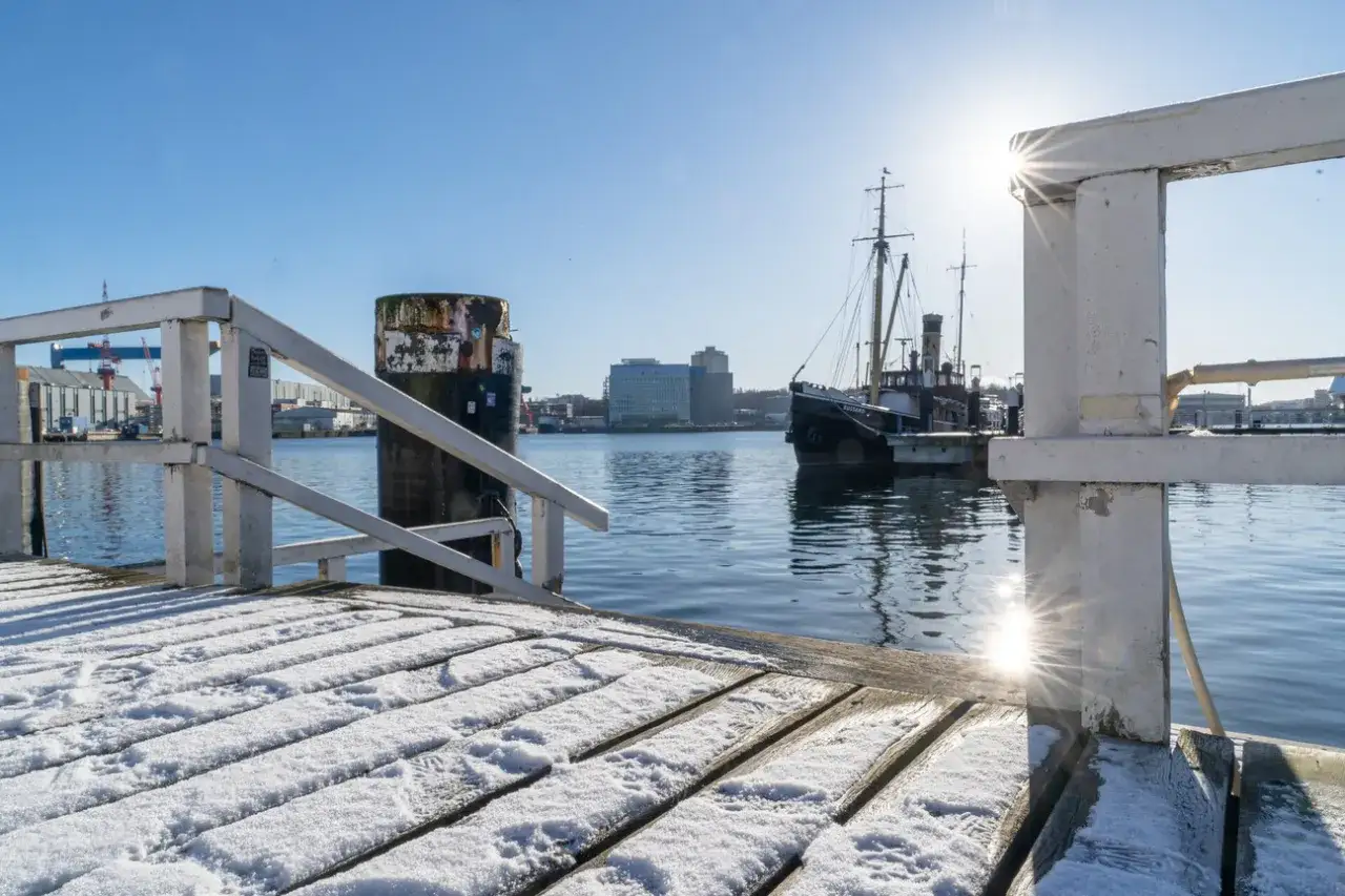Kiel im Winter: Ein schneebedecktes Holzdeck f&uuml;hrt zu einem alten Schiff im Hafen. Die Sonne blendet.