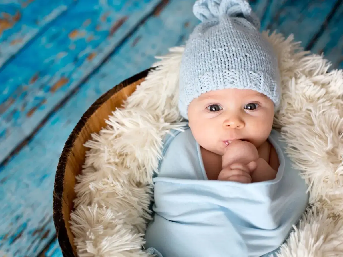 Bebé con gorro azul, envuelto en manta, en una cesta con peluche. Su nombre, Gala, significa