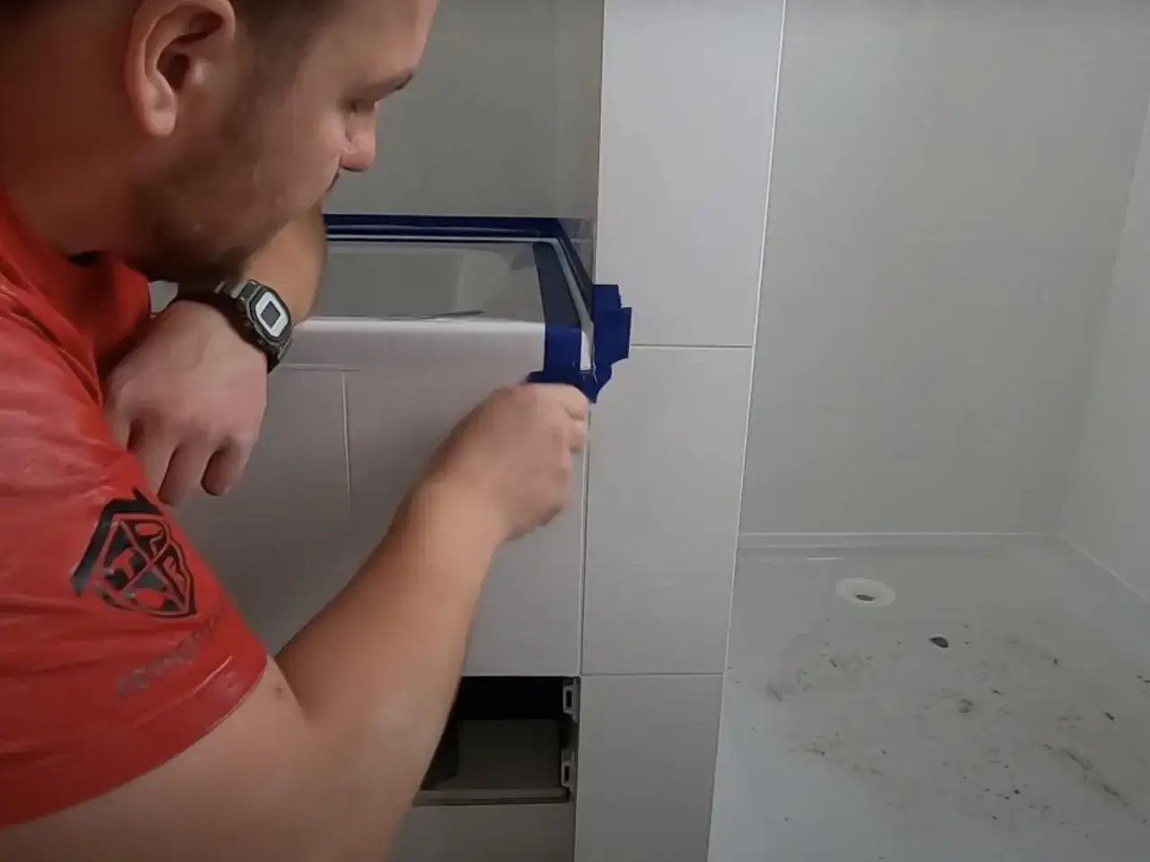 Mężczyzna w czerwonej koszulce przygotowuje się do silikonowania płytek, zaklejając krawędzie taśmą.