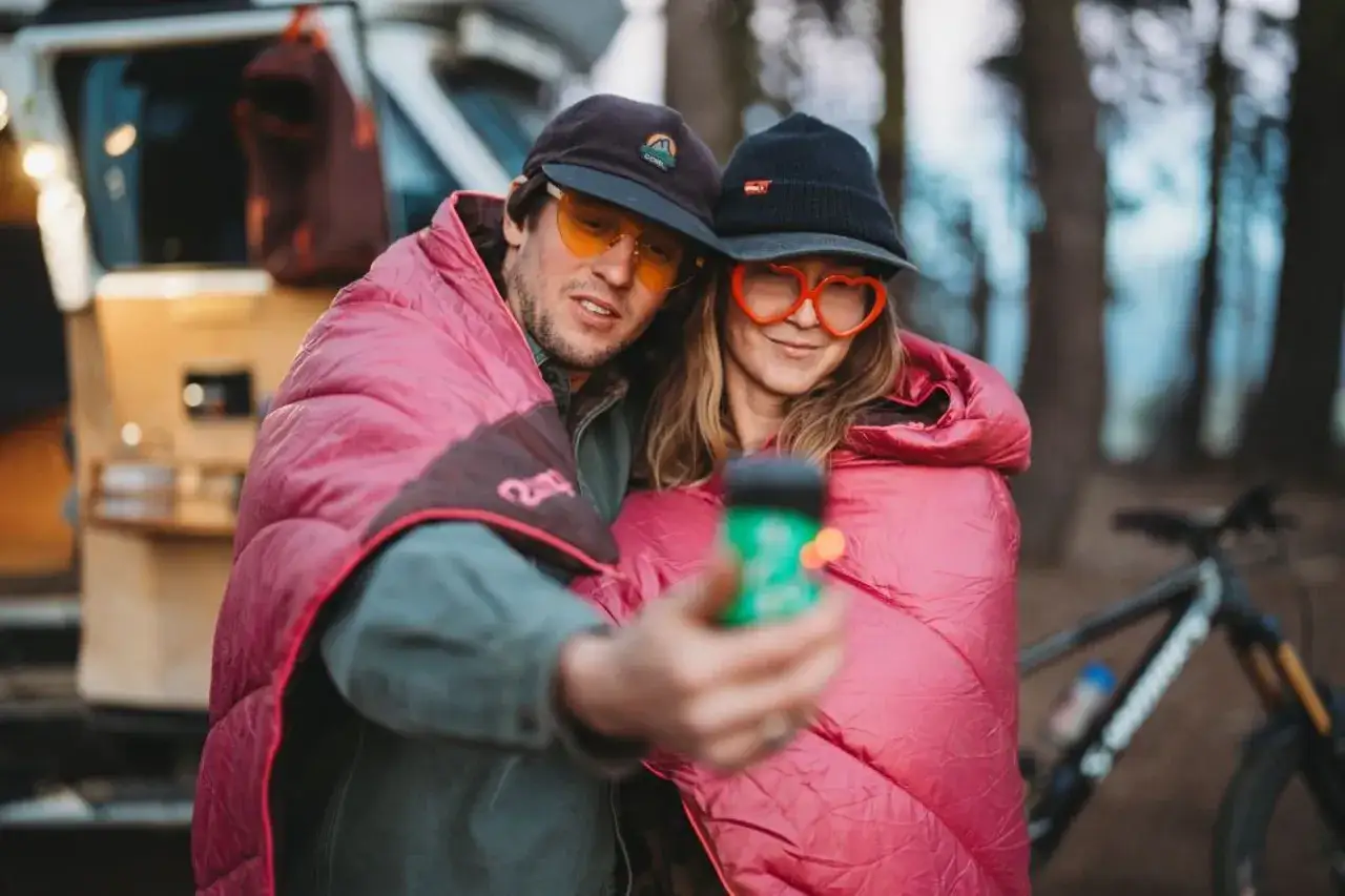 Ein Paar im Freizeithaus, eingewickelt in rosa Decken, macht ein Selfie. Sie tragen lustige Brillen und Mützen. Perfekter Freizeitpark-Outfit!