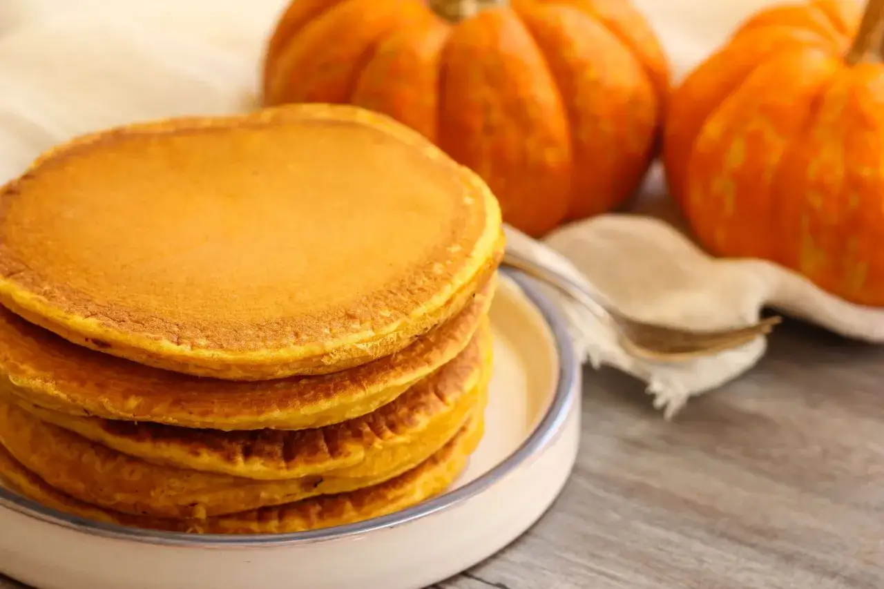 Torrijas de calabaza doradas y esponjosas apiladas en un plato, con calabazas decorativas al fondo.