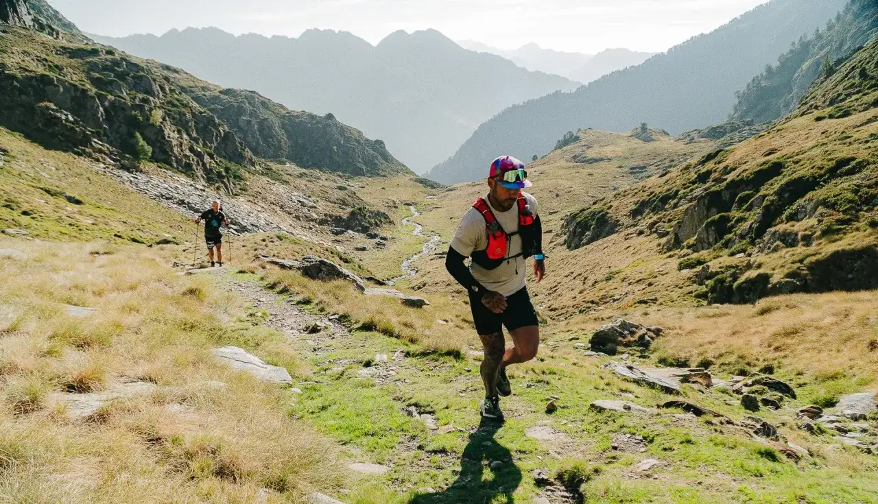 Deux coureurs s'attaquent au **transpyrénéenne trail** dans un paysage montagneux spectaculaire, entre herbes sèches et rochers.