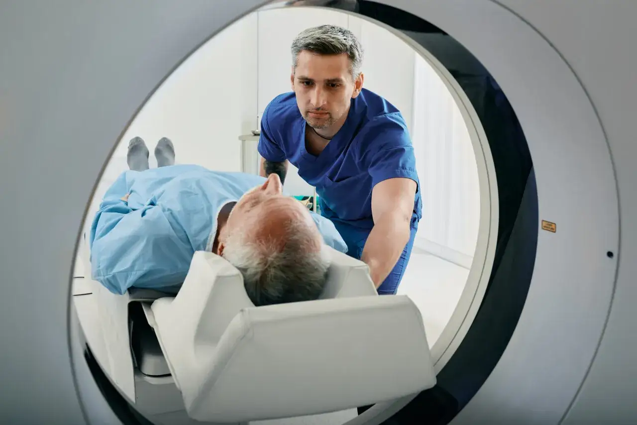 Pacjent w szpitalnej piżamie leży na stole tomografu. Technik medyczny pomaga mu, przygotowując do badania. Nie trzeba się rozbierać do tomografii.
