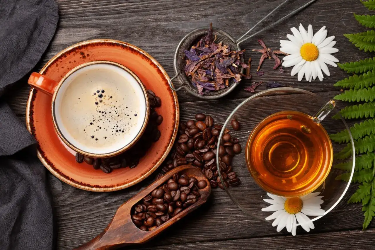 Une tasse de caf&eacute; cr&eacute;meux et une infusion de th&eacute; ambr&eacute;, accompagn&eacute;es de grains de caf&eacute;, de fleurs s&eacute;ch&eacute;es et de camomille sur fond de bois.