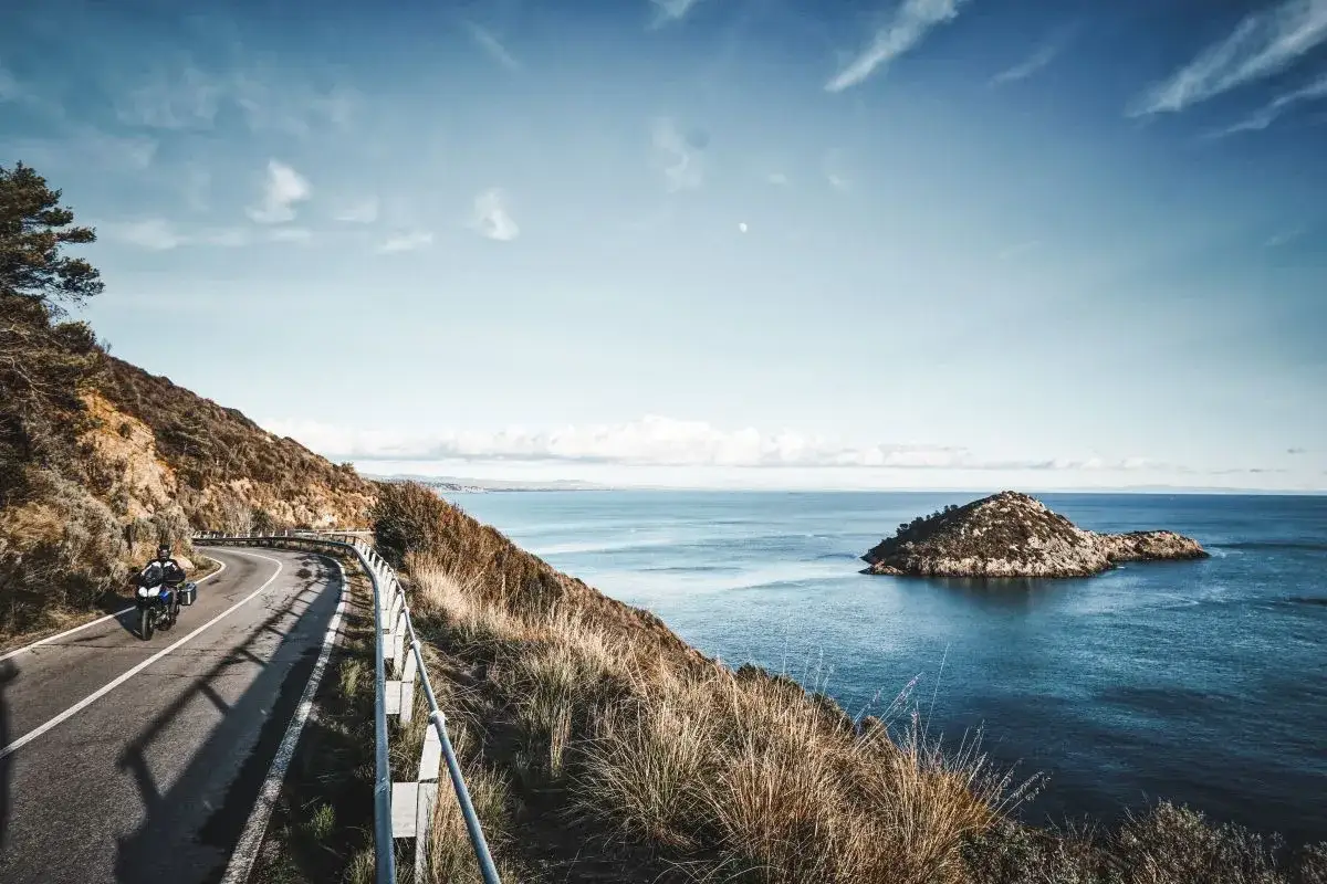 Motociclista percorre la strada panoramica verso Sperlonga, con il mare blu e un isolotto roccioso.