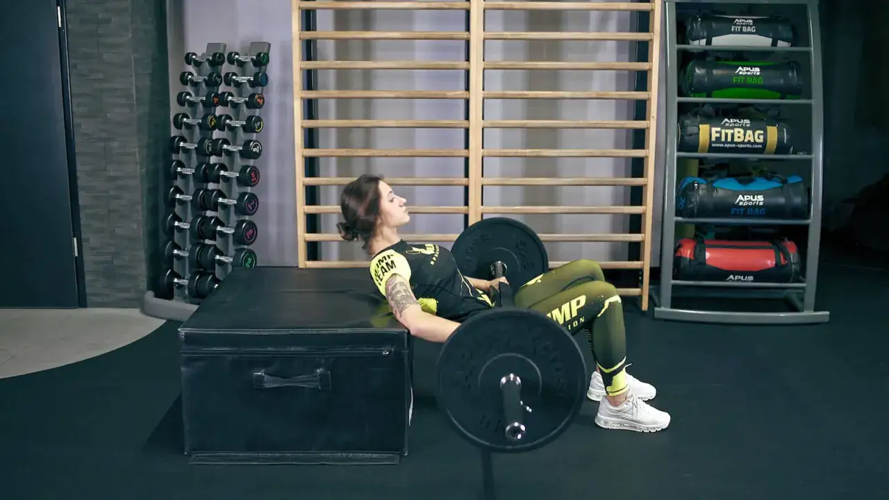 Kobieta wykonuje ćwiczenie hip thrust z ciężarem na ławce. W tle stojaki z hantlami i torbami treningowymi.