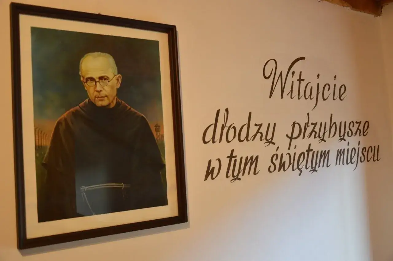 Obraz przedstawia portret błogosławionego Maksymiliana Kolbe w habicie, z widokiem na obóz koncentracyjny w tle. Obok napis: