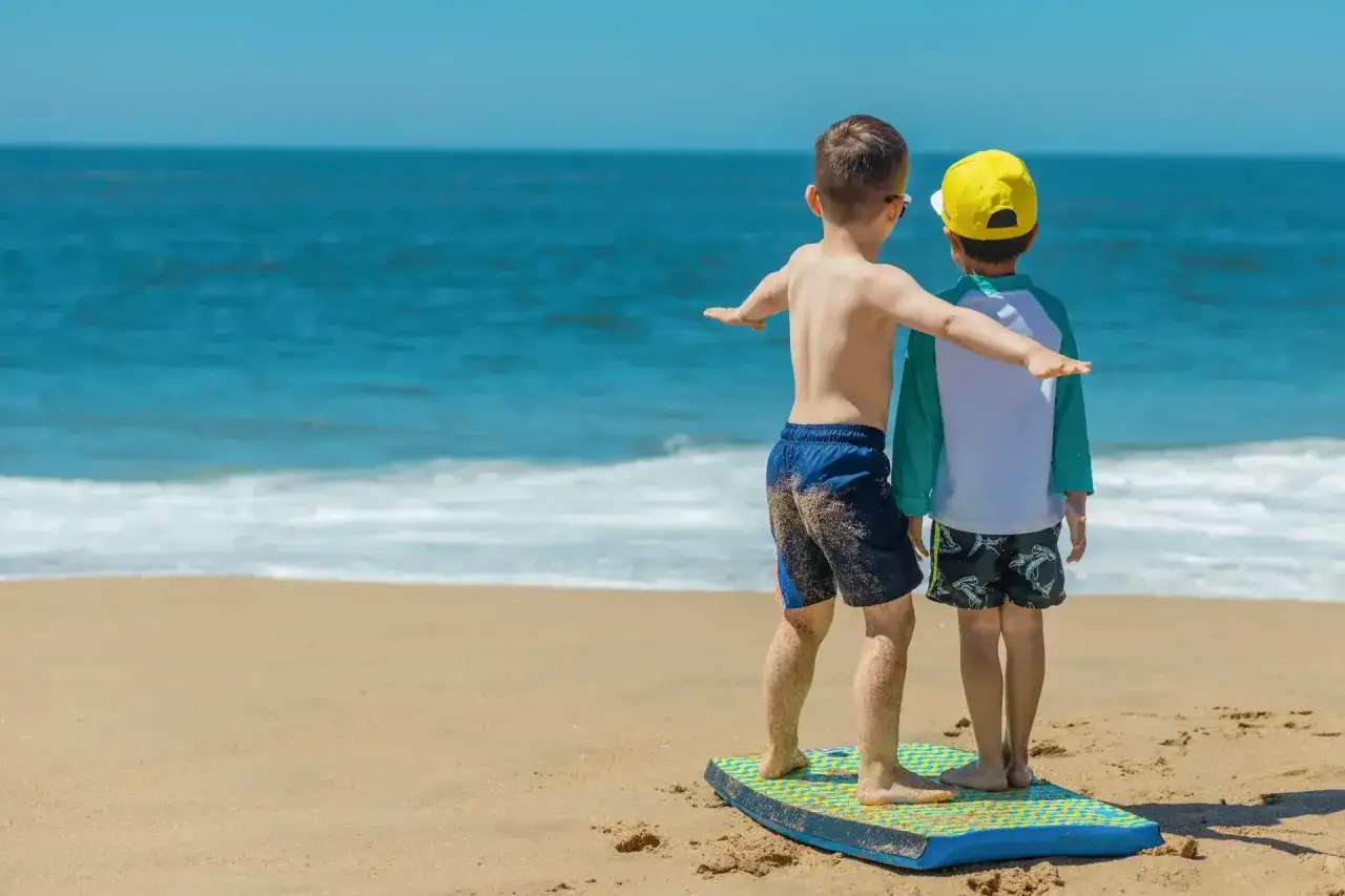 Dwóch chłopców na desce surfingowej na plaży, cieszących się wakacjami z dziećmi za granicą.