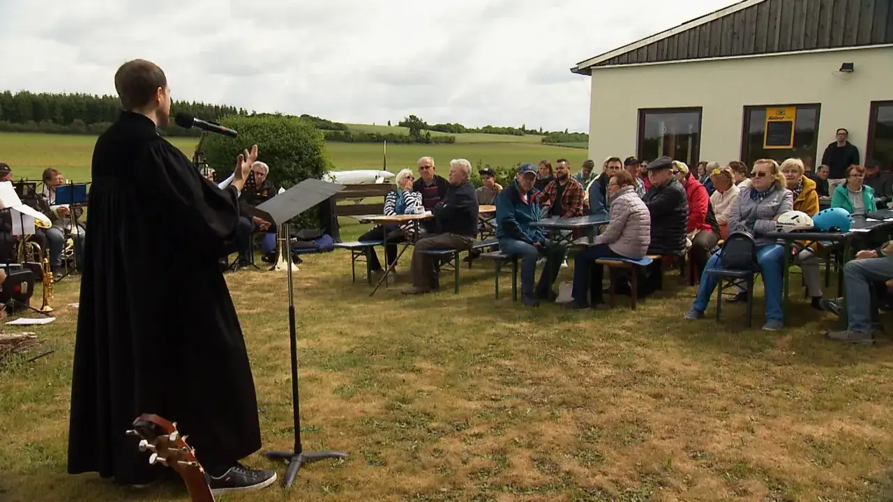 Feierliche Feier zu Christi Himmelfahrt im Freien. Ein Geistlicher spricht zu einer versammelten Gemeinde, während eine Blaskapelle im Hintergrund spielt.