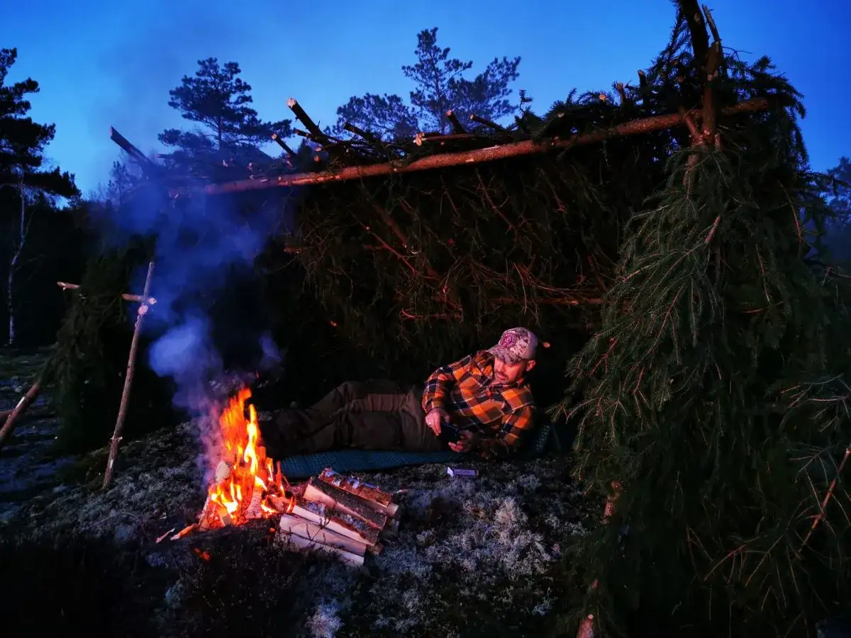 Man vilar vid en eld under ett provisoriskt vindskydd byggt av granris.