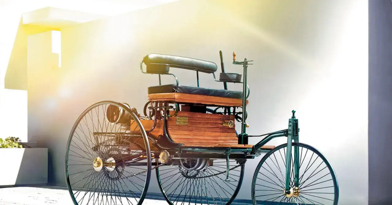 Pierwsze auto świata, Benz Patent-Motorwagen, z drewnianą karoserią i wielkimi szprychowymi kołami, stoi na tle jasnej ściany.