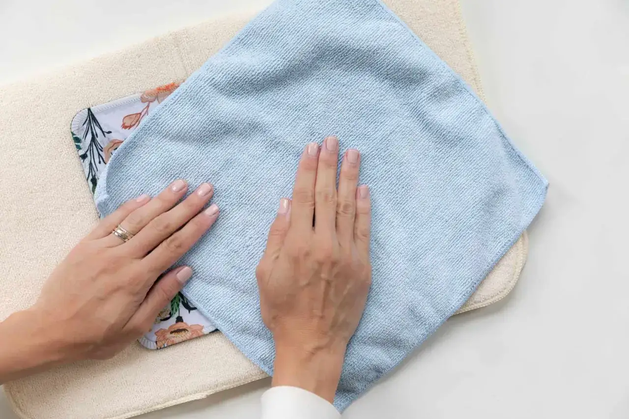 Hände reinigen ein bunt gemustertes Mauspad mit einem hellblauen Mikrofasertuch.