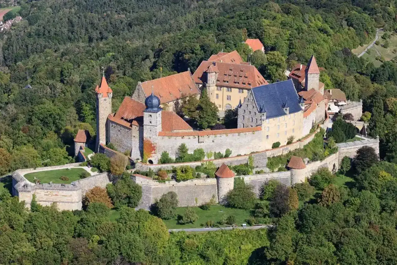 Die Veste Coburg thront über der Coburg Altstadt, umgeben von grünen Wäldern. Eine imposante Burganlage mit Türmen und Mauern.
