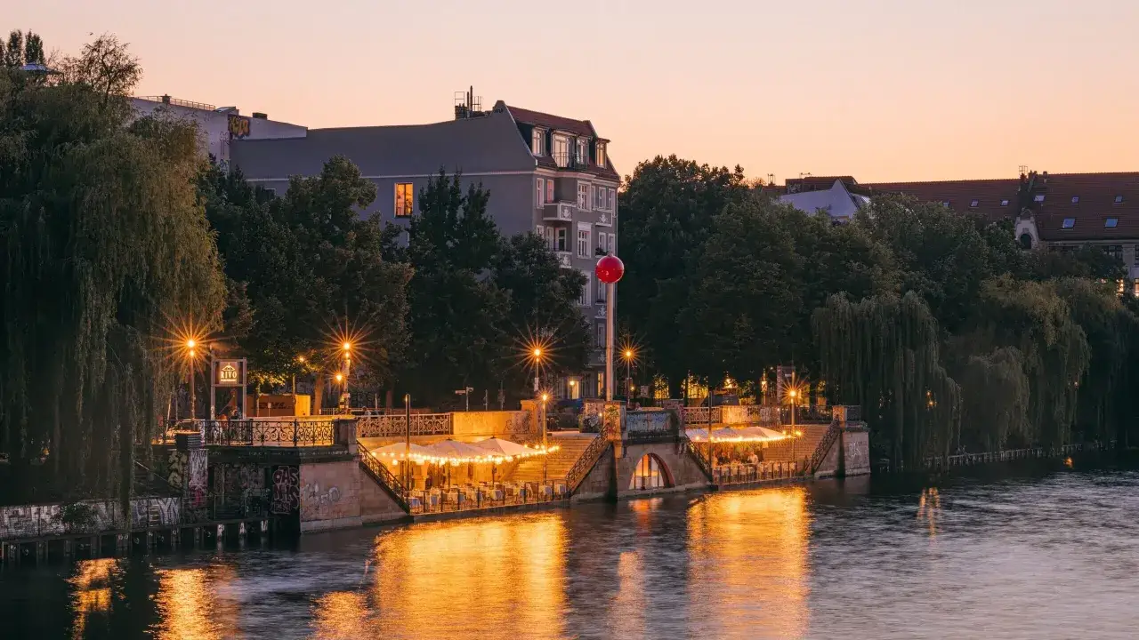Abendstimmung an einem Restaurant am Wasser. Lichterketten beleuchten Tische unter Sonnenschirmen, w&auml;hrend die Spiegelung im Fluss gl&auml;nzt.
