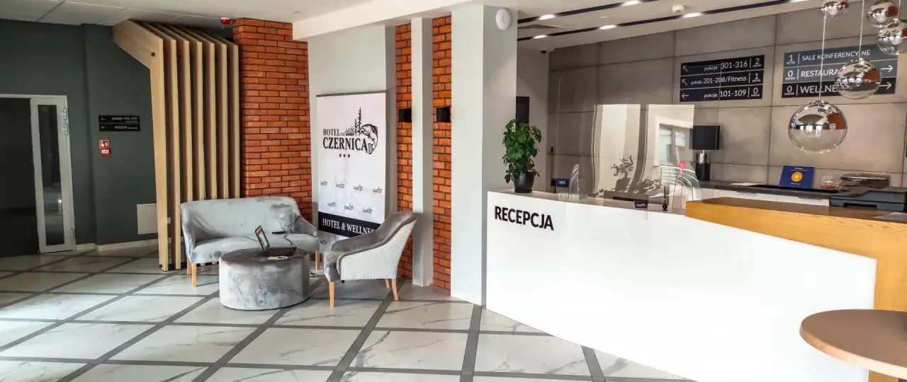 Recepcja hotelu Czernica. Wygodne fotele i stolik z laptopem, obok baner hotelu. Znaki kierunkowe sugerują, że może być overbooking, bo pokoje są wszędzie.