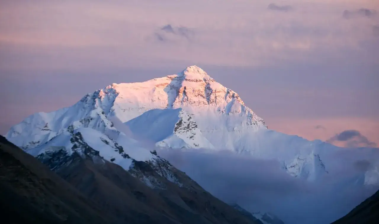 Ośnieżony szczyt Mount Everest w blasku wschodzącego słońca. Widać skaliste zbocza i chmury otulające górę.