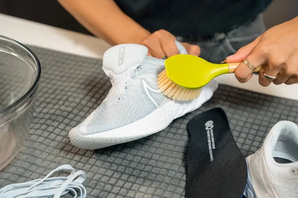 Hände reinigen mit einer Bürste die weißen Mesh Schuhe. Daneben liegen Schnürsenkel und eine Einlegesohle.