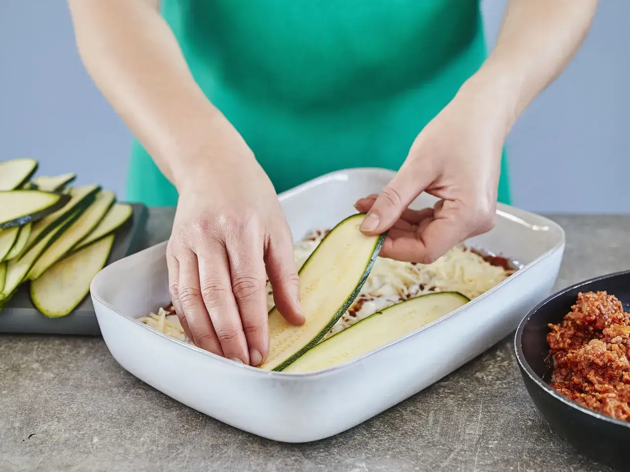 H&auml;nde legen Zucchinischeiben auf eine Schicht K&auml;se und Hackfleisch in einer Auflaufform. Zucchini-Lasagne mit Lasagneplatten wird zubereitet.