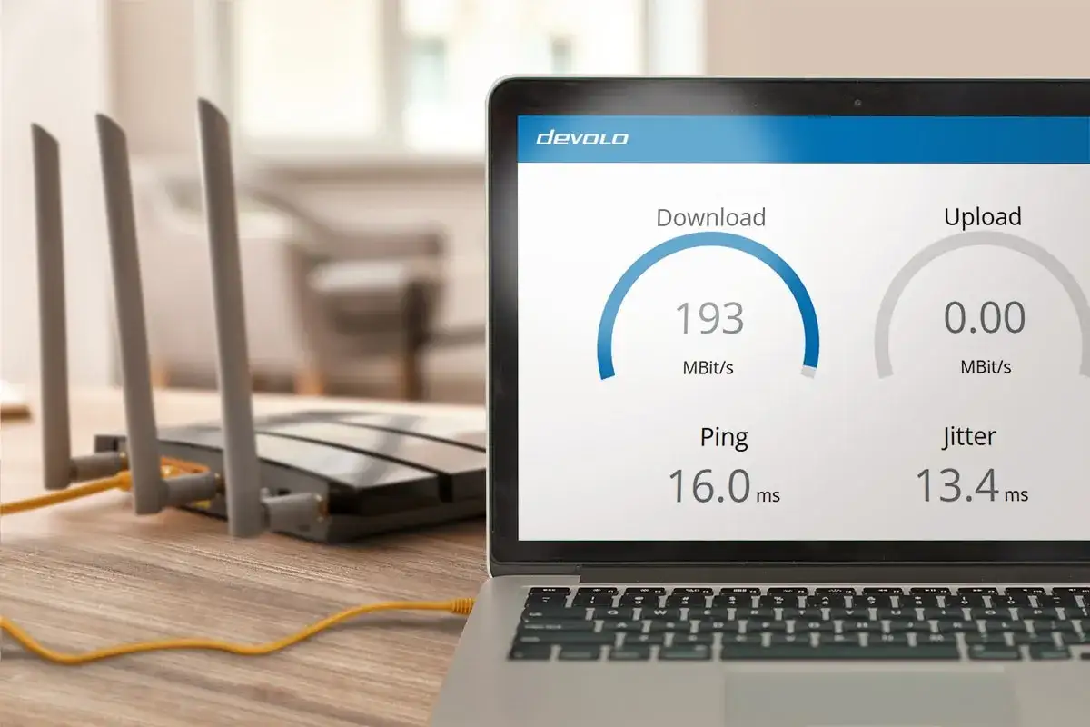 Laptop zeigt 193 MBit/s Download, 0.00 MBit/s Upload, Ping 16.0 ms und Jitter 13.4 ms. Ein WLAN-Router steht daneben.