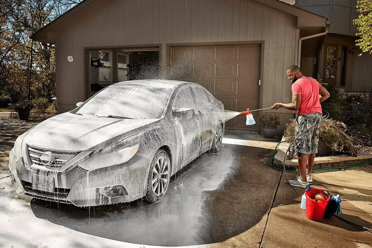 Hombre aplicando espuma a un coche gris para lavarlo en casa. Cubo rojo y manguera cerca.