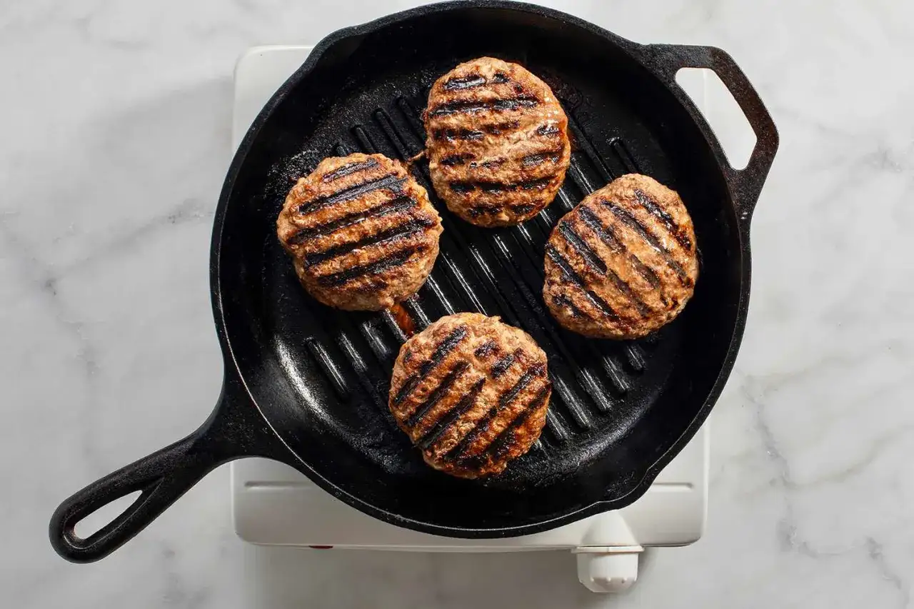 Vier saftige Burger-Patties brutzeln in einer Gusseisenpfanne und entwickeln k&ouml;stliche Grillstreifen.
