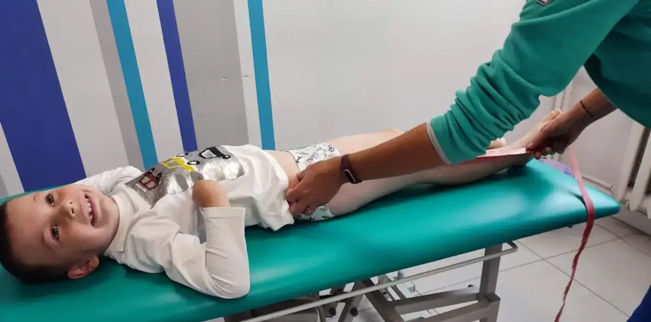 Chłopiec leży na kozetce, a fizjoterapeuta mierzy mu nogi. Dziecko uśmiecha się, choć jedna noga kr&oacute;tsza od drugiej wymaga ćwiczeń.