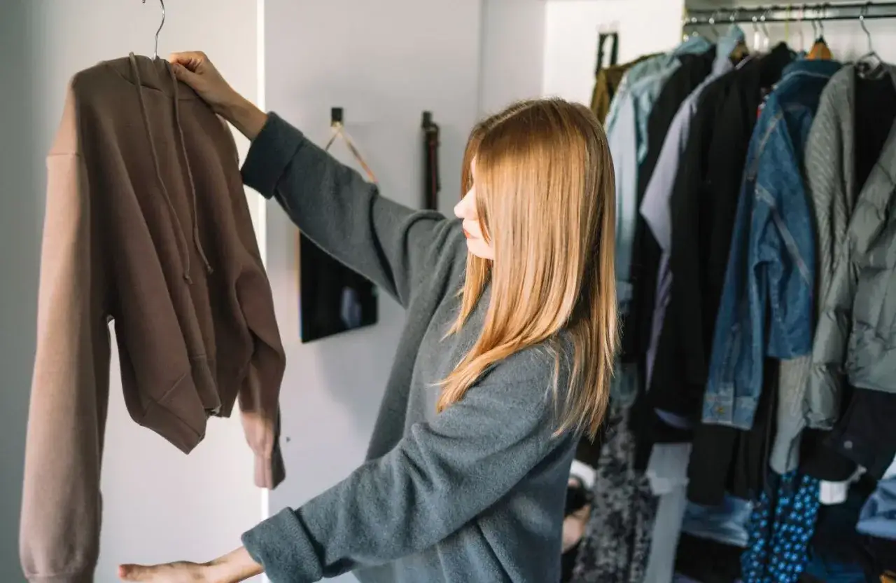 Une femme examine un pull marron, réfléchissant à comment trier ses vêtements. Son dressing est bien rangé.
