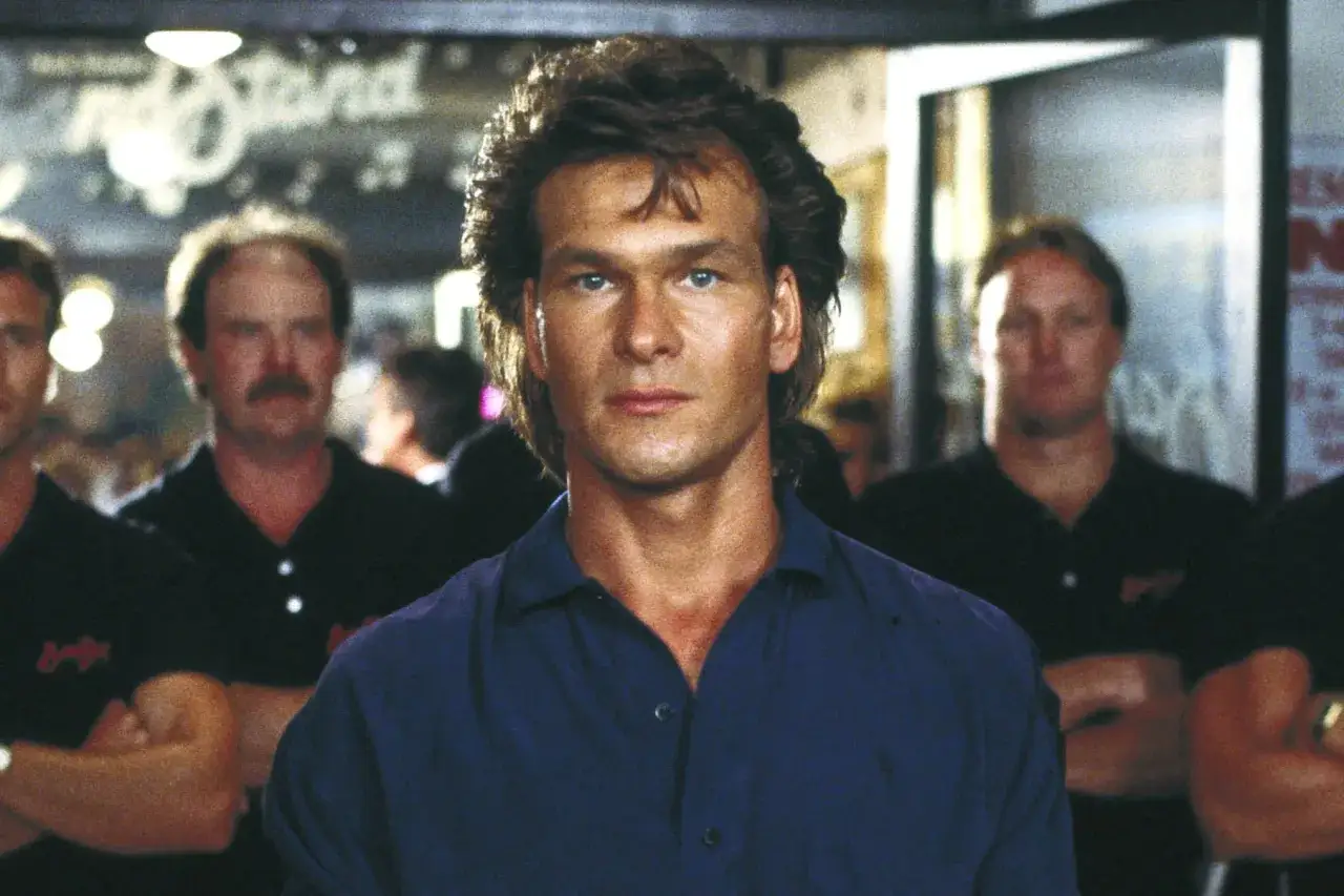 Patrick Swayze w filmach, z charakterystyczną fryzurą i niebieską koszulą, otoczony przez innych mężczyzn.