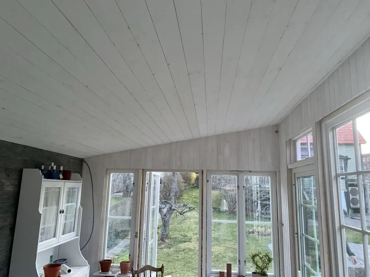 Taket och väggarna är målade med vit råspont. Ett vitt skåp står till vänster, och stora fönster ger utsikt över en grönskande trädgård.