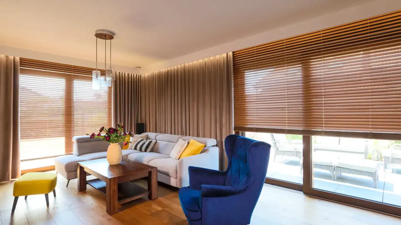 Elegancki salon z szarym narożnikiem, granatowym fotelem i żółtym pufem. Okna zdobią **żaluzje drewniane i zasłony**, tworząc przytulną atmosferę.