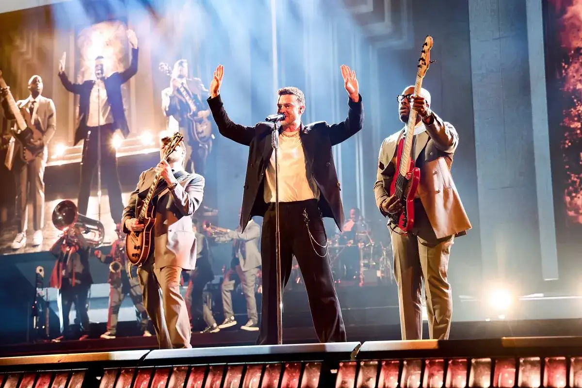 Justin Timberlake w Polsce na koncercie, śpiewa z rękami w g&oacute;rze, otoczony przez muzyk&oacute;w grających na gitarach i instrumentach dętych.