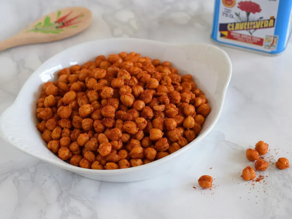 Un taz&oacute;n blanco lleno de garbanzos fritos crujientes y especiados. Una lata de especias y una cuchara de madera decorada est&aacute;n al fondo.