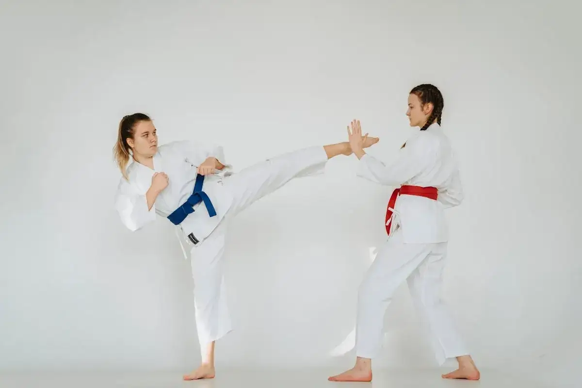 Två kvinnor i vita dräkter tränar kenpo karate. Den ena utför en hög spark, den andra blockerar.
