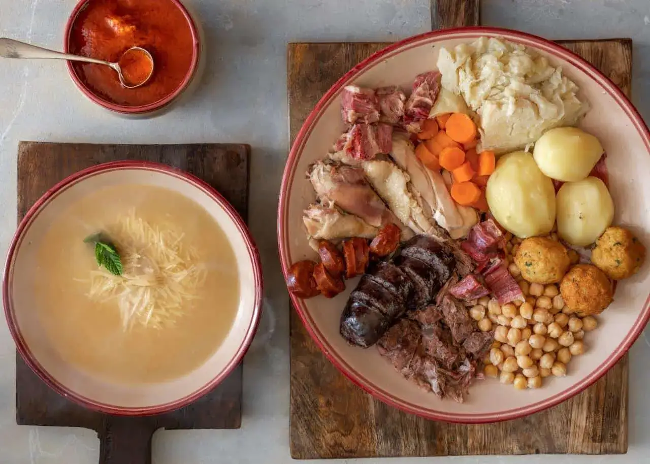 Un plato generoso de cocido madrile&ntilde;o con garbanzos, carnes variadas, chorizo, morcilla, patatas, zanahorias y repollo, acompa&ntilde;ado de sopa de fideos y salsa de tomate.
