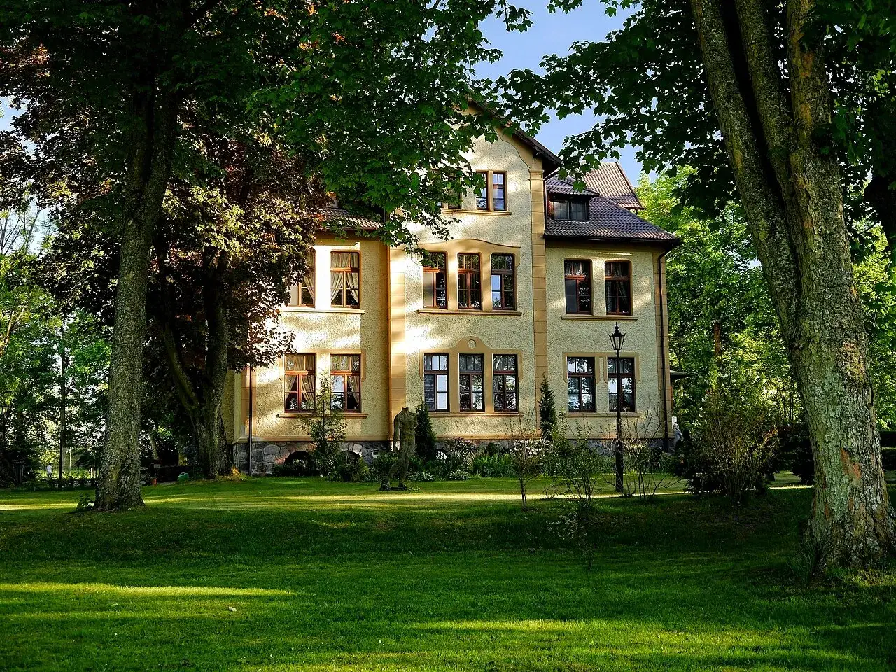 Jasny pałac Poraj otoczony zielenią, z trawnikiem i drzewami w słoneczny dzień.