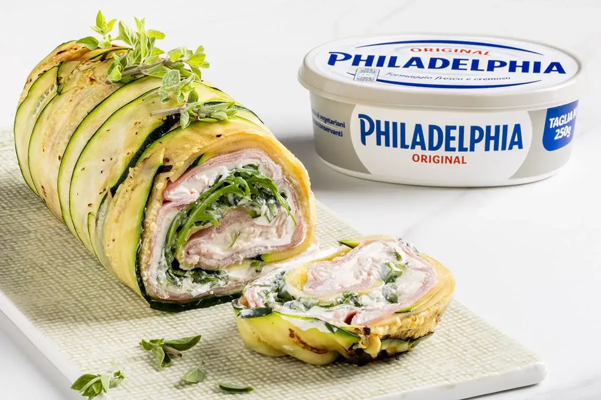 Rotolo di zucchine ripieno di prosciutto e formaggio fresco, guarnito con erbe aromatiche. Accanto, una confezione di Philadelphia.