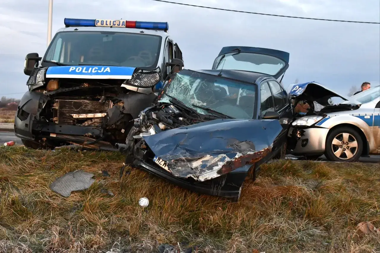 Zniszczone samochody po wypadku, w tym radiow&oacute;z. Widoczny prz&oacute;d ciemnego auta, maska wygięta, szyba pęknięta. Przepadek pojazdu po kolizji.
