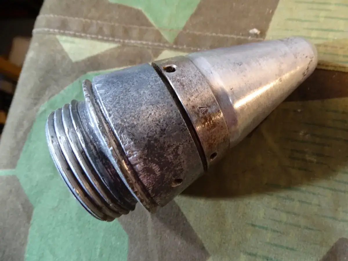 Metallisches Teil einer Flak Granate mit Gewinde und spitz zulaufendem Ende, liegt auf Tarnstoff.