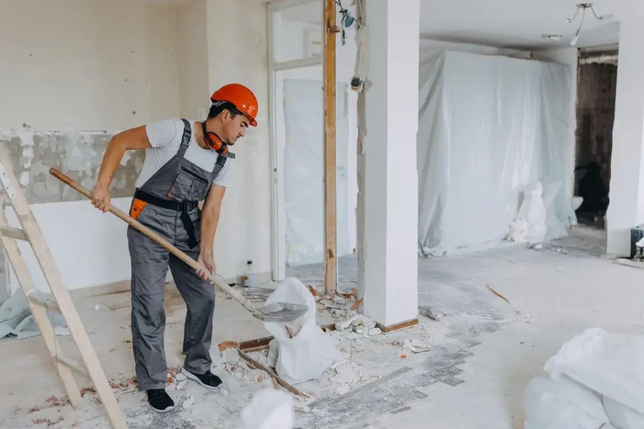 Un ouvrier en casque orange pelle des d&eacute;bris. La poussi&egrave;re de pl&acirc;tre est omnipr&eacute;sente, un danger &agrave; ne pas n&eacute;gliger lors de ces travaux.