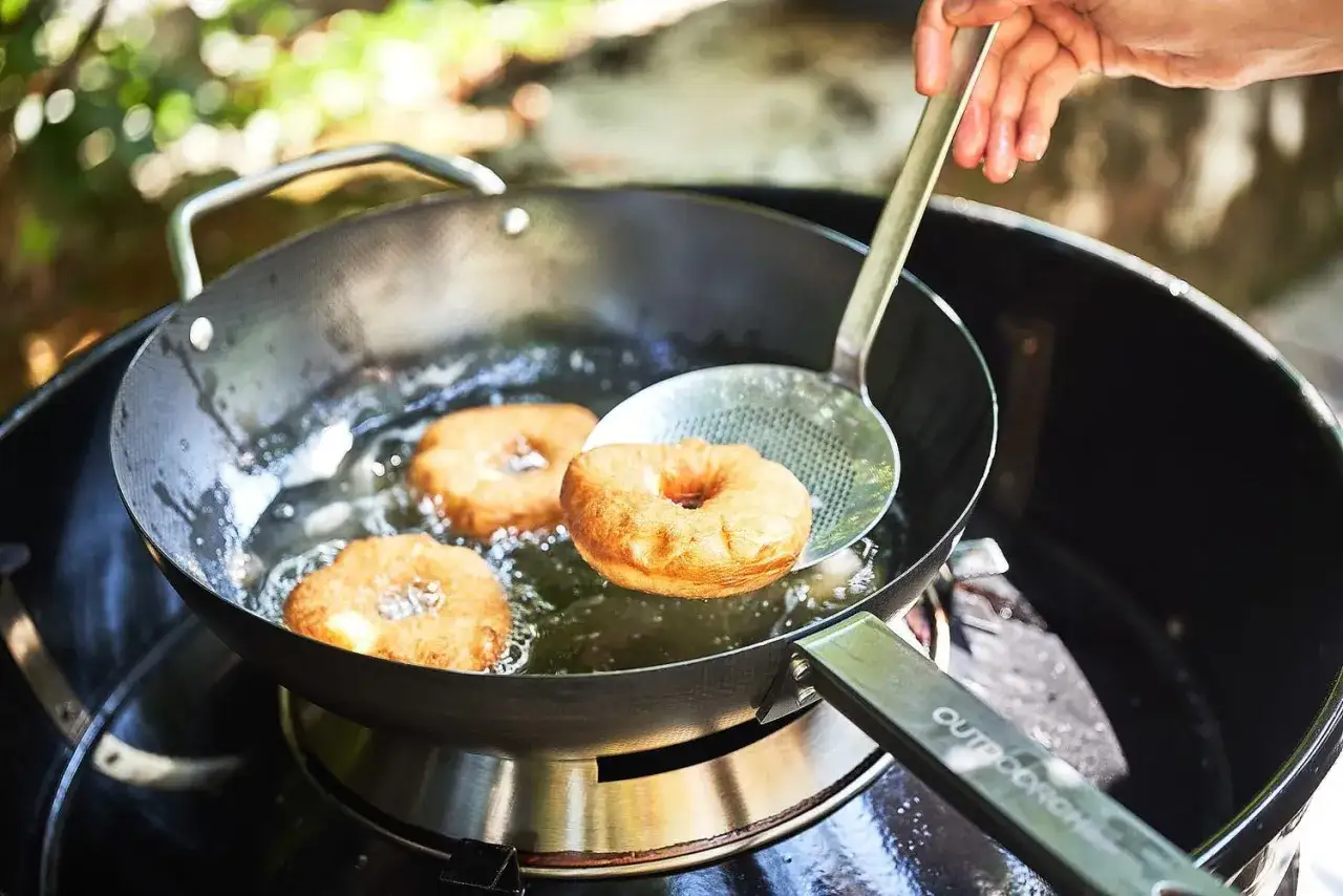 Friture de beignets dorés dans un wok sur un barbecue. Une main utilise une écumoire pour retirer un beignet de l'huile chaude.