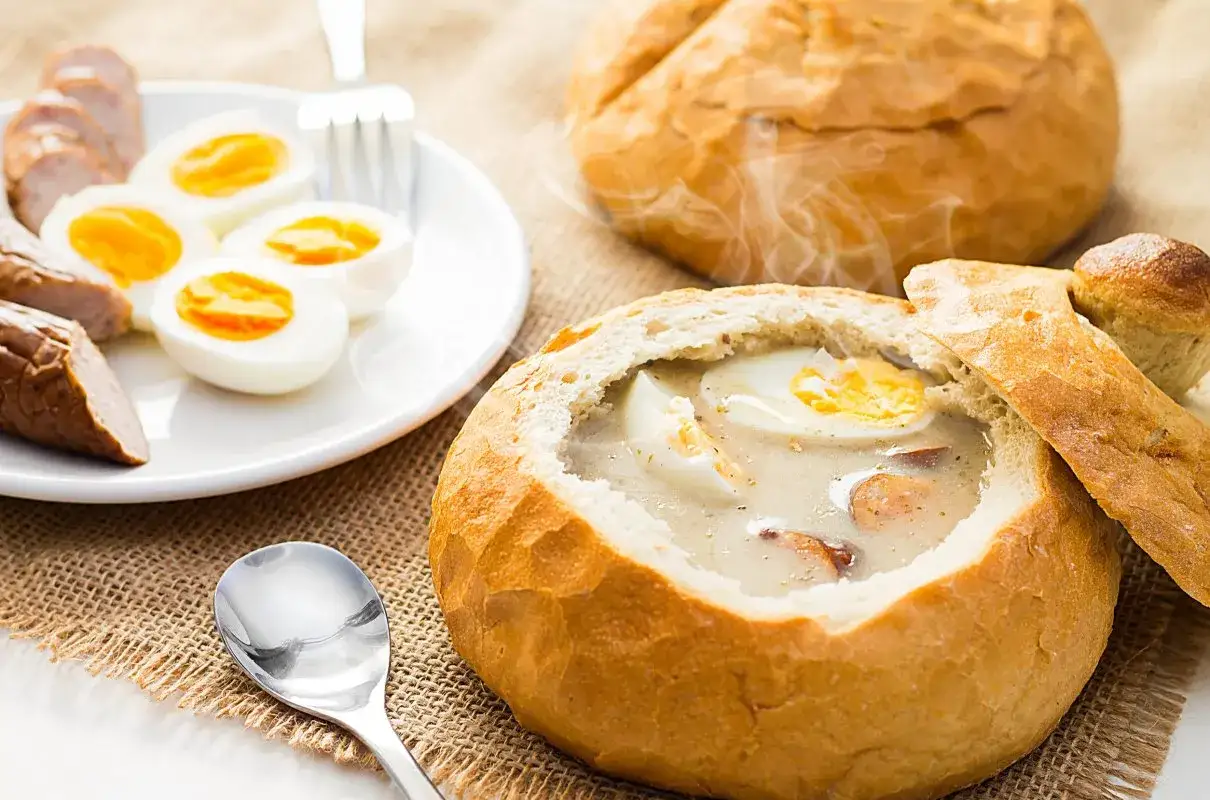 Gorący żurek w chlebie, podany z jajkiem i kiełbasą, paruje apetycznie. Obok talerz z połówkami jajek i kawałkami kiełbasy.
