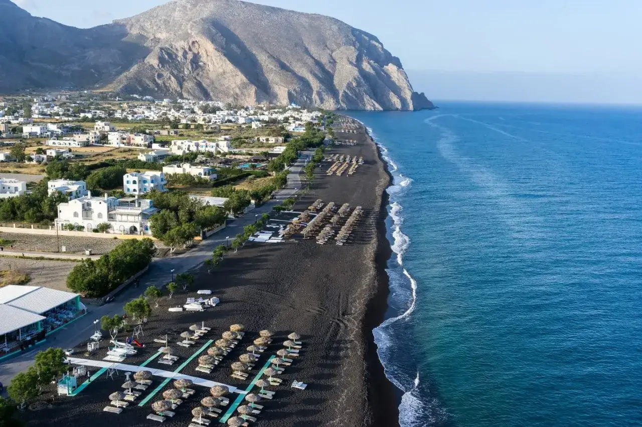 Ein atemberaubender schwarzer santorini strand mit Liegen und Sonnenschirmen, daneben weiße Häuser und ein bergiger Hintergrund.