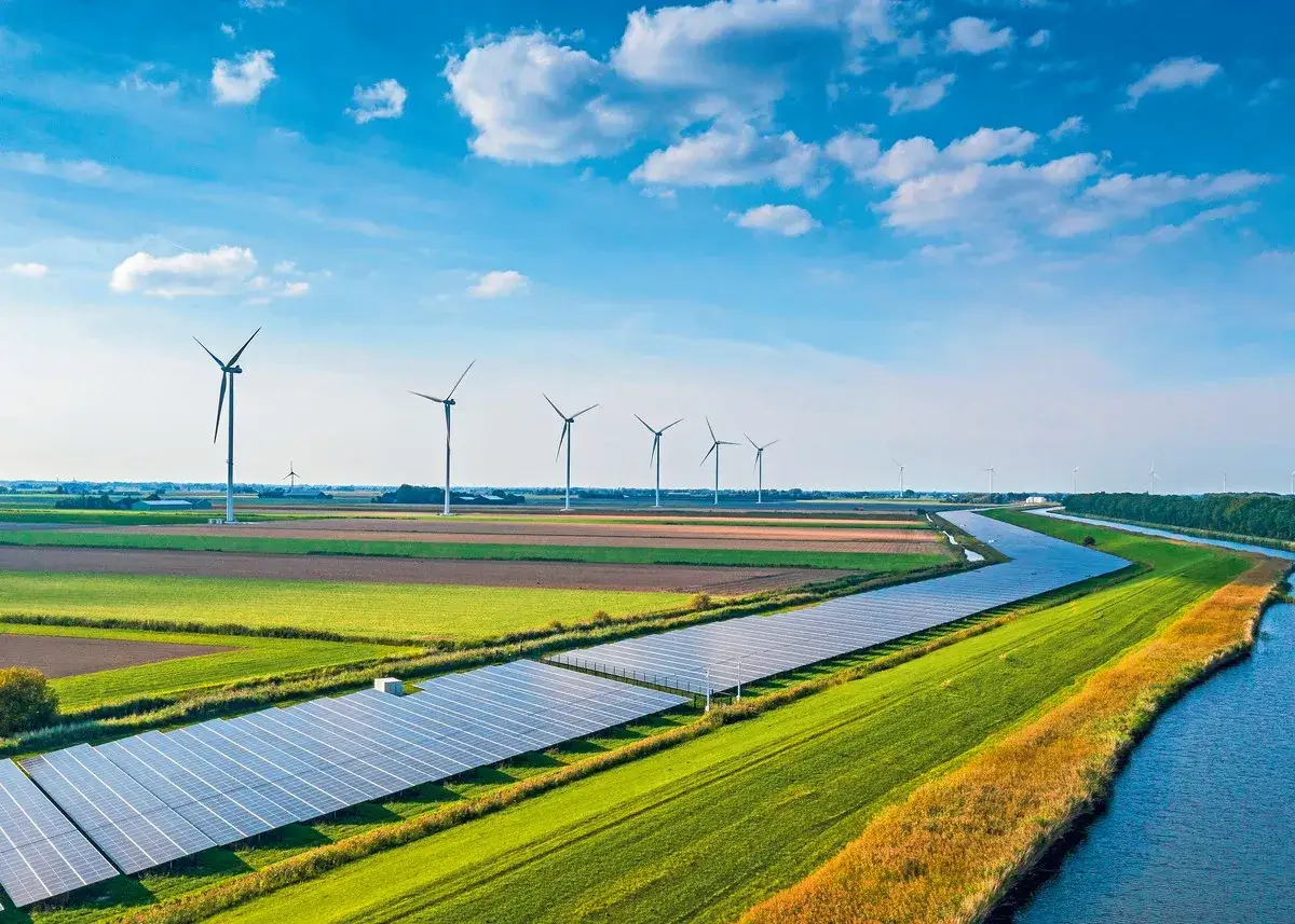 Wiatraki energetyczne i panele słoneczne na tle błękitnego nieba z chmurami. Zielone pola i kanał wodny tworzą krajobraz zrównoważonej energii.