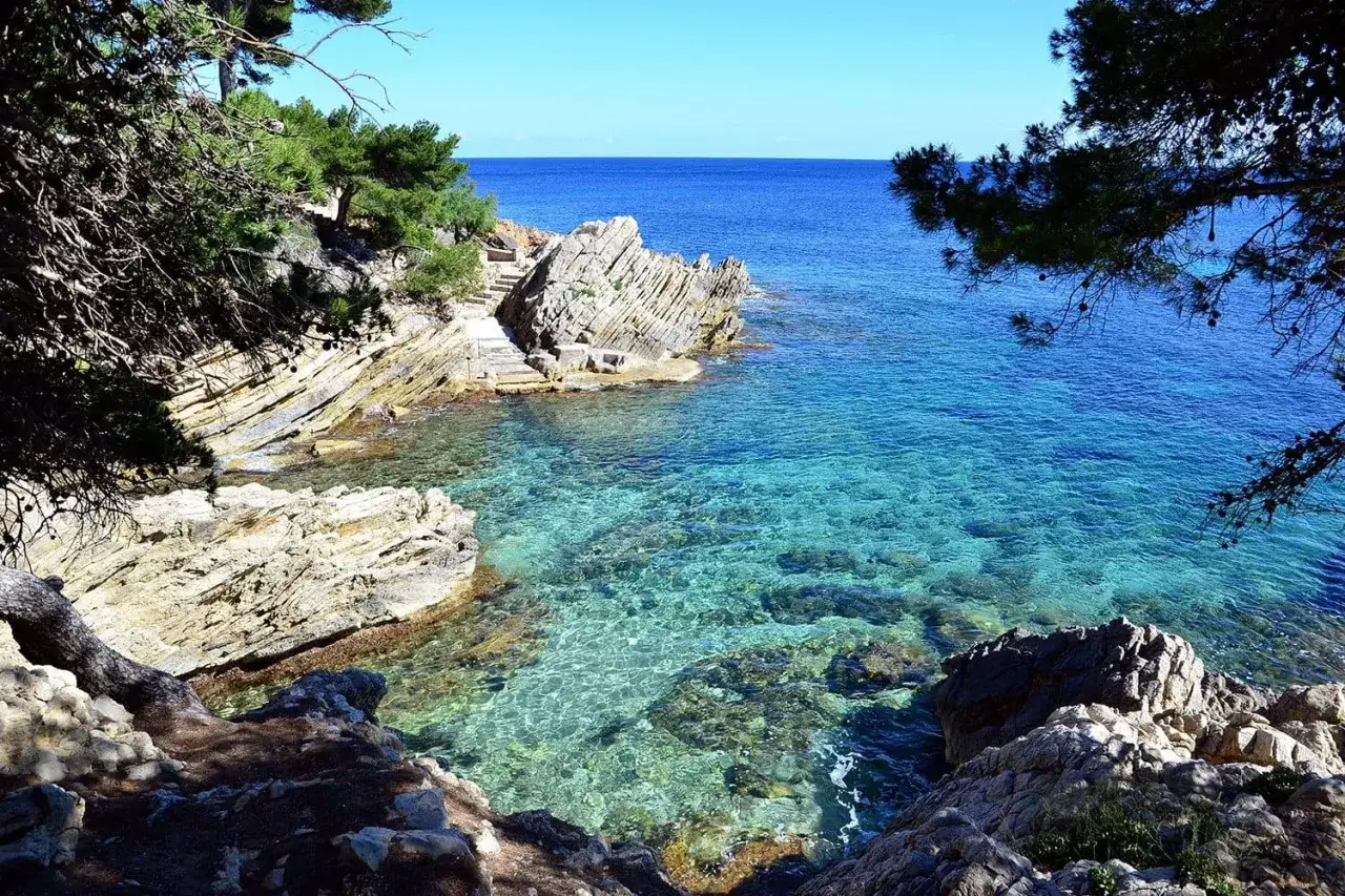 Agua cristalina en una cala rocosa, con escaleras de piedra que bajan al mar. El sol ilumina el fondo marino, invitando a explorar esta cala gata.