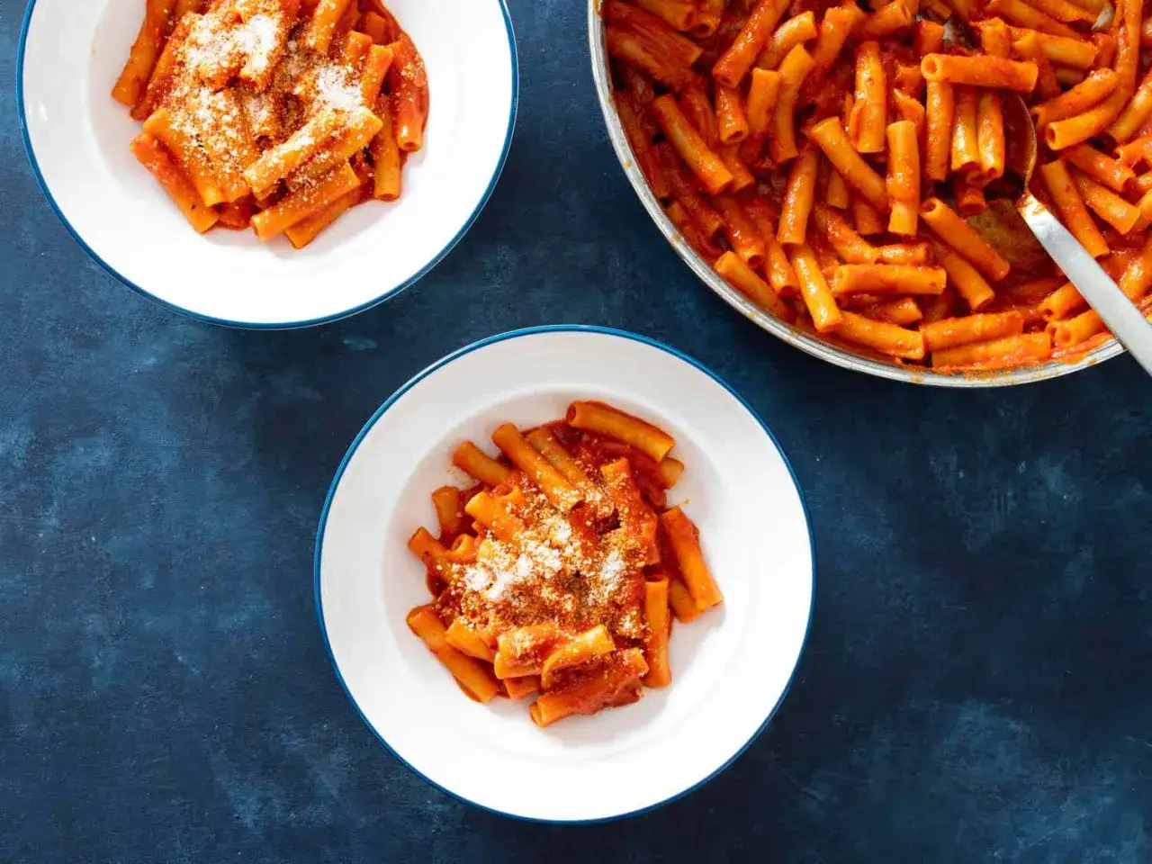 Två tallrikar med rörformad pasta i en krämig tomatsås, toppad med riven ost. En panna med mer nduja pasta syns i bakgrunden.