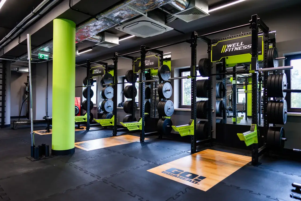 Siłownia z wolnymi ciężarami i rackami. Jasnozielona kolumna i logo 