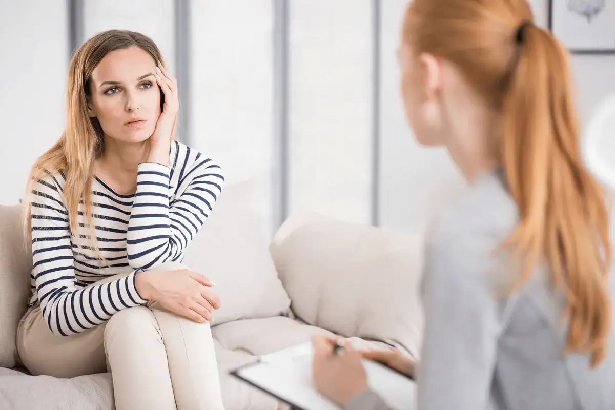 Kobieta siedzi na kanapie, zamyślona, podczas sesji terapeutycznej. Zadaje pytania do psychologa, szukając wsparcia.